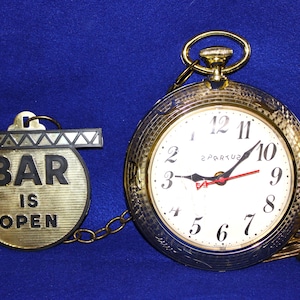 May include: A gold-toned pocket watch-style wall clock with a white face, black numerals, and the brand name Spartus. A gold chain connects the clock to a circular metal sign with black text that reads BAR IS OPEN, featuring a decorative top bar.