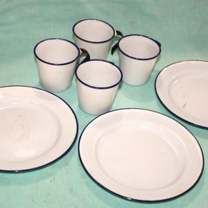 May include: A white enamelware dish set with a blue trim. The set includes four mugs and three plates. The mugs have black handles. The plates and mugs are arranged on a light green surface.