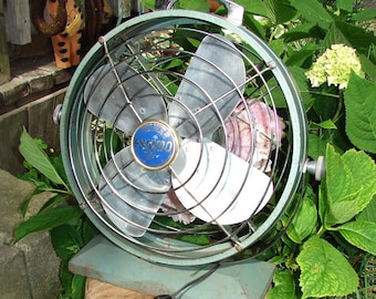 May include: A vintage green metal fan with a white and silver blade. The fan has a blue and white label that says "Eskimo" on it. The fan is sitting on a wooden base and is surrounded by green plants.