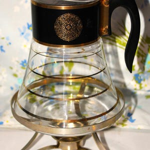 May include: Vintage glass coffee carafe with a black top and handle, accented with gold trim and a decorative medallion. The carafe sits on a silver-toned metal stand with three legs. Horizontal gold bands encircle the glass body.
