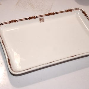 May include: Rectangular white ceramic serving dish with a brown decorative border. The border features a repeating floral design and a small emblem in the center. The dish has a slightly distressed appearance.