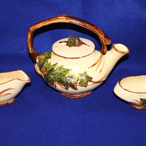 May include: A cream-colored ceramic tea set consisting of a teapot with a brown branch-like handle and lid, accompanied by a matching creamer and sugar bowl. Each piece features raised green ivy leaf motifs and brown vine accents on the exterior.