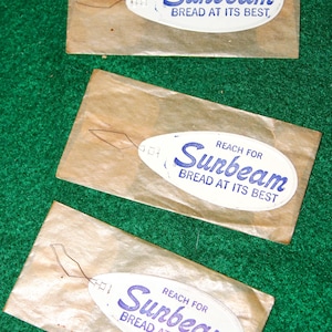 May include: Three rectangular paper packets, each containing a metal needle threader with a white oval tag. The tag features blue text that reads REACH FOR Sunbeam BREAD AT ITS BEST. The threaders are attached to the paper with thin wire loops.