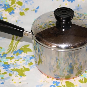 May include: A stainless steel saucepan with a lid and a black handle. The pot has a copper-colored bottom and a black lid handle. The background is a floral pattern with blue and white flowers.