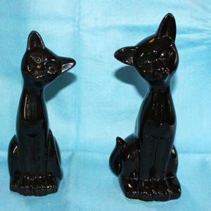 May include: Two glossy black ceramic cat figurines in a seated position. Both cats feature elongated necks, pointed ears, and smooth, reflective surfaces. The cat on the left is tilted slightly to the side, while the cat on the right faces forward.