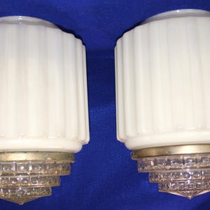May include: A pair of cylindrical white milk glass light shades featuring vertical ribbing. Each shade is attached to a tiered, clear glass base with a square grid pattern and a metallic ring connector at the junction of the two glass sections.