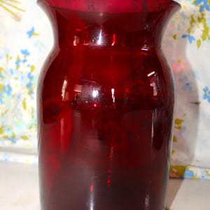 May include: A deep red glass vase with a wide body and flared rim. The vase is made of transparent glass, allowing light to pass through. The vase is likely used for decorative purposes.