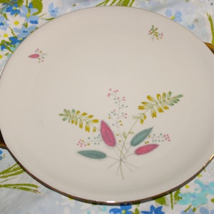 May include: A round, white ceramic serving plate with gold trim and handles. The plate features a floral design with pink, yellow, and teal leaves and sprigs. The plate is set against a floral patterned background.
