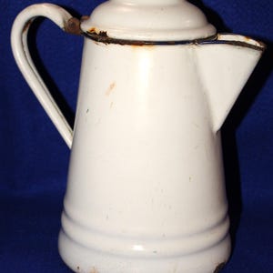 May include: A white enamel coffee pot with a matching lid, a curved handle, and a wide spout. The metal surface shows visible signs of wear, including rust along the rim, the base, and the handle attachment point, with a smooth, glossy finish.