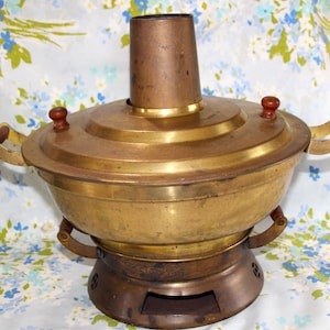 May include: A vintage brass hot pot with a copper chimney and wooden handles. The pot has a round base and a lid with two wooden knobs. The handles are made of brass and wood. The pot is sitting on a floral patterned background.