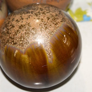 May include: A close-up of a ceramic sphere with a speckled brown top and a glossy, dark brown base. The sphere has a unique glaze with hints of gold and purple, creating a textured, artistic appearance. The background is blurred.