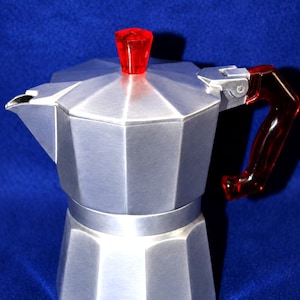 May include: A silver-colored aluminum moka pot featuring an octagonal body, a hinged lid with a faceted red translucent knob, and a matching red translucent handle. The coffee maker has a metallic finish and a prominent spout on the side.