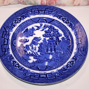 May include: A round, blue and white ceramic plate with a Willow pattern design. The plate features a central scene of a pagoda, trees, and a bridge, surrounded by a decorative border. The plate is likely for decorative or dining use.