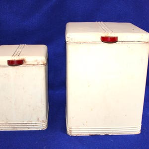 May include: Two rectangular off-white metal canisters of different heights, each featuring a hinged lid with a small red handle and horizontal ribbed detailing at the base and top. The containers have a smooth finish with visible signs of wear.