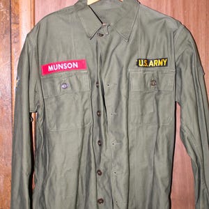 May include: An olive green long-sleeve button-down shirt featuring a red name patch with the text MUNSON on the right chest and a black patch with the yellow text U.S. ARMY on the left chest. The shirt has two front pockets with button closures.