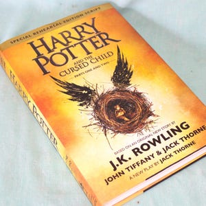 May include: Hardcover book titled "Harry Potter and the Cursed Child." The cover is predominantly orange with black text and a graphic of a nest with wings. The author's name, J.K. Rowling, is also visible.