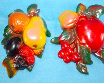 May include: Two three-dimensional wall plaques shaped like clusters of fruit. The left plaque features an orange, a yellow pear, a dark plum, and red and black berries. The right plaque displays a peach, a large red apple, and red grapes.
