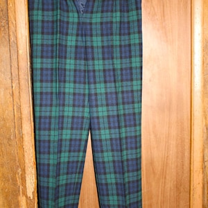 May include: Green, navy, and black plaid pants hanging on a silver hanger. The pants have a button and zipper closure. The plaid pattern is a classic design, suitable for various occasions. The pants are made of a woven fabric.