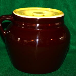May include: A dark brown ceramic crock with a glossy finish, featuring a single side handle and a round, flat lid with a bright yellow top surface and a white rim. The container has a wide, rounded body and a short, narrow neck opening.