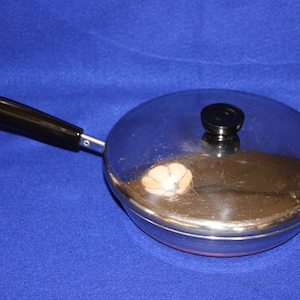 May include: A round stainless steel skillet with a polished finish and a matching lid. The pan features a long, black heat-resistant handle with a small metal hanging loop at the end. A black knob is centered on the lid for easy lifting.