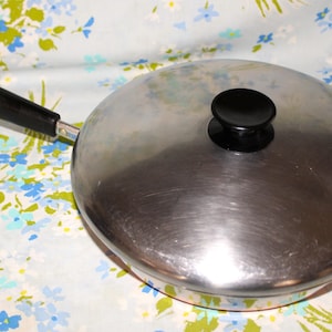 May include: A stainless steel frying pan with a lid. The pan has a black handle and a black knob on the lid. The pan is sitting on a floral patterned fabric with blue, white, and green flowers.