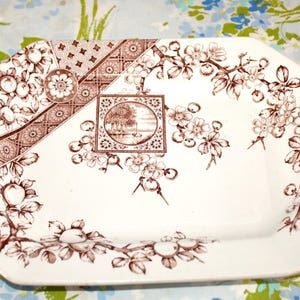 May include: An octagonal white serving platter with a brown floral and landscape design. The platter features a central square landscape scene and floral border. The design includes flowers, leaves, and fruit.