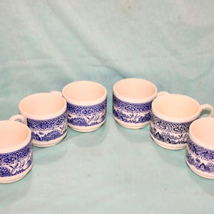 May include: Six white ceramic mugs with blue decorative patterns. Each mug has a handle and a blue and white design around the exterior. The mugs are arranged on a light blue surface.