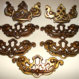May include: A collection of seven antique brass drawer pulls. The pulls have an ornate, scrolled design with a warm, golden finish. The hardware is likely for furniture restoration or decorative use.