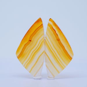 Puede incluir: Dos rodajas de ágata pulidas en tonos naranja y crema. Las piedras muestran un patrón bandeado, con una calidad translúcida. Las formas son aproximadamente triangulares y se presentan sobre un fondo blanco. El ágata es probablemente de origen natural.