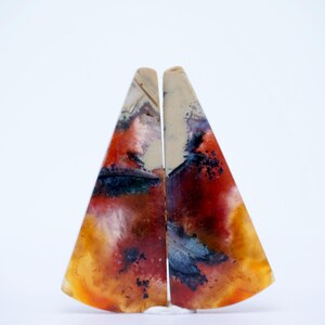 Può includere: Due pietre preziose lucide di forma triangolare. Le pietre mostrano un vivace mix di colori, tra cui rosso, arancione, giallo, blu e crema. I motivi assomigliano a paesaggi astratti. Le pietre sono esposte su sfondo bianco.