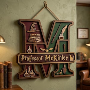 May include: A wooden wall hanging shaped like the letter M, featuring a maroon and green background. It displays a wizard hat, books, a wand, a quill, potions, a golden snitch, and a broom. A horizontal plaque reads Professor McKinley.