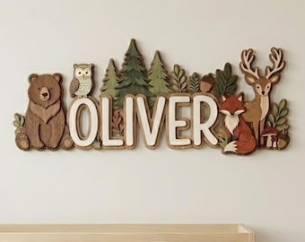 Könnte beinhalten: Holz-Wanddekoration für das Kinderzimmer mit dem Namen "OLIVER" in weißen Buchstaben, umgeben von Waldtieren und Bäumen. Zu den Tieren gehören ein Bär, eine Eule, ein Fuchs und ein Hirsch. Die Dekoration befindet sich über einem Kinderbett.