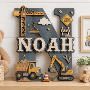 May include: A large wooden letter N wall decoration in dark blue, featuring the name NOAH in cream-coloured letters. The piece is decorated with yellow construction vehicles, including a crane, a dump truck, and an excavator, plus a yellow sign reading LITTLE BUILDER, a traffic cone, stars, and a cloud.