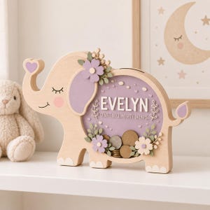 Può includere: Un salvadanaio in legno chiaro a forma di elefante con un pannello frontale in acrilico trasparente. Il design presenta un orecchio viola, un cuore sulla proboscide e fiori. Il testo recita EVELYN e GENTLE SOUL, MIGHTY DREAMS. Sono visibili monete.