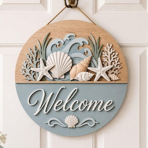 May include: A circular wooden wall sign featuring a natural wood top half with layered cutouts of blue waves, white starfish, and various seashells. The bottom half is painted dusty blue with the white script text Welcome and a small shell icon.