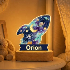 May include: A rocket-shaped night light featuring a watercolor space design with stars, a crescent moon, a planet, and a galaxy. The base is made of light-colored wood, and a dark blue rectangular plaque at the bottom displays the name Orion in white.