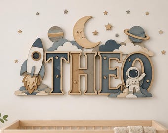 May include: A wooden wall decoration featuring the name THEO in large block letters, surrounded by space-themed elements including a rocket ship, a crescent moon with a face, planets, clouds, and small stars in shades of blue, tan, and white.