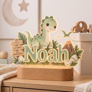 Puede incluir: Un letrero luminoso de acr&iacute;lico con un dinosaurio verde de dibujos animados, un volc&aacute;n y huevos. El nombre Noah aparece en letras grandes de color verde claro. Un poste de madera dice Little Explorer Big Adventures. La pieza descansa sobre una base de madera.