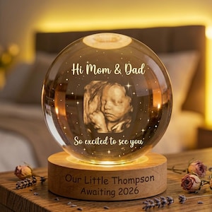 Può includere: Una sfera di cristallo luminosa con un'immagine 3D di un bambino, appoggiata su una base di legno. La sfera riporta il testo "Hi Mom & Dad" e "So excited to see you". La base recita "Our Little Thompson Awaiting 2026".