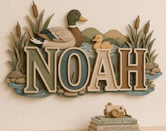 May include: A wall-mounted wooden sign featuring the name NOAH in large, multi-colored block letters. The design includes a mallard duck, a duckling, cattails, and rolling hills. Below, a small wooden toy camera sits on a stack of three books.