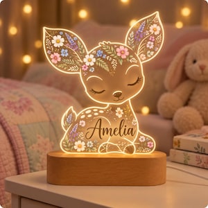May include: A light-up acrylic lamp shaped like a fawn with closed eyes, featuring a floral pattern in pink, white, and blue. The name Amelia is printed in black script on the fawn's body. The lamp sits on a smooth, oval-shaped wooden base.