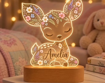 May include: A light-up acrylic lamp shaped like a fawn with closed eyes, featuring a floral pattern in pink, white, and blue. The name Amelia is printed in black script on the fawn's body. The lamp sits on a smooth, oval-shaped wooden base.