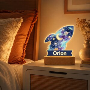 May include: A rocket-shaped night light featuring a watercolor space design with stars, a crescent moon, a planet, and a galaxy. The base is made of light-colored wood, and a dark blue banner at the bottom displays the name Orion in white text.