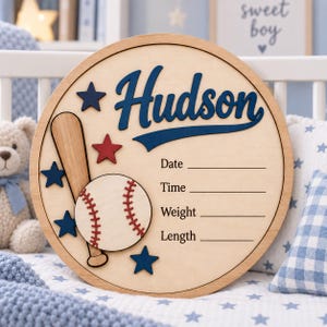 Könnte beinhalten: Ein rundes Geburtsanzeigeschild aus Holz mit dem Namen Hudson in blauer Schreibschrift, einem Baseballschläger, einem Baseball mit roten Nähten und mehreren blauen und roten Sternen. Das Schild enthält Zeilen für Datum, Uhrzeit, Gewicht und Länge.