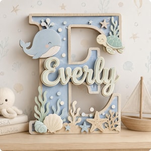 May include: Wooden letter "E" with a light blue background and sea-themed decorations. Features a whale, turtle, starfish, and the name "Everly" in white script. Decorative elements include seashells and coral.