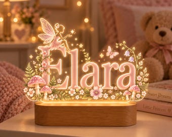May include: A custom illuminated acrylic sign featuring the name Elara in soft pink lettering. The design includes a fairy silhouette, butterflies, small flowers, and mushrooms, all mounted on a rectangular wooden base that emits a warm glow.