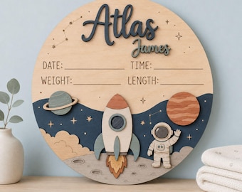 Puede incluir: Un letrero circular de madera para anunciar nacimientos con temática espacial, que incluye un cohete, un astronauta, planetas y estrellas. El letrero tiene el nombre Atlas James en relieve y líneas para fecha, hora, peso y longitud.