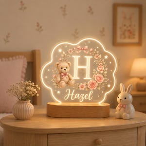 May include: A cloud-shaped acrylic night light with a wooden base, featuring an illuminated letter H, a teddy bear, and a floral wreath. The name Hazel is printed in script below the bear. The light glows with a warm, soft white radiance.