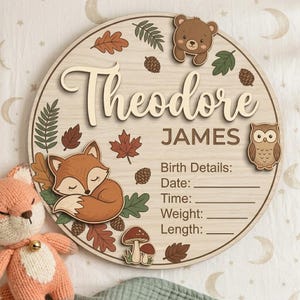 May include: A wooden round sign with the name "Theodore James" in a script font, surrounded by woodland-themed illustrations. Includes a sleeping fox, bear, owl, leaves, and birth details section.
