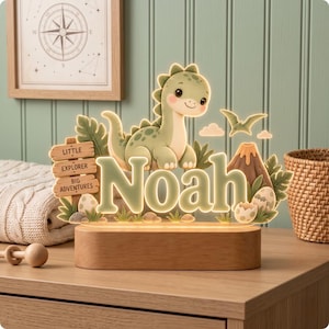 Puede incluir: Un letrero luminoso de acr&iacute;lico con un dinosaurio de dibujos animados, un volc&aacute;n y el nombre Noah en letras grandes. El letrero incluye una base de madera y un peque&ntilde;o poste con el texto Little Explorer Big Adventures, rodeado de follaje.